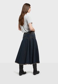 Falda maxi de mezclilla en azul oscuro, diseño acampanado con dobladillo deshilachado. Combinada con una camiseta gris de manga corta y botas negras hasta la rodilla.