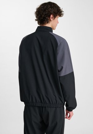 Zwarte en grijze jas met een opstaande kraag, elastische tailleband en contrasterende mouwen. Gladde, lichte stof met een relaxte pasvorm.