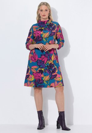 Blumendruckkleid mit blauem Hintergrund, ausgestattet mit pinken, lila und gelben Blättern. 3/4-Ärmel, hoher Kragen und ausgestellt gesäumt. Kombiniert mit lila Stiefeletten.