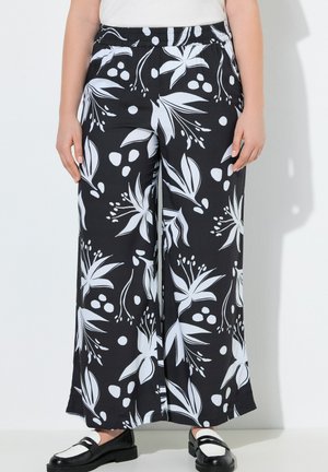 Kvinde iført sorte bukser med brede ben med stort hvidt blomsterprint og sorte-hvide loafers, stående mod en ensfarvet lys baggrund.