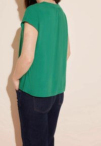 Donna che indossa una blusa verde a maniche corte e jeans blu scuro, in piedi con le mani nelle tasche su uno sfondo beige.