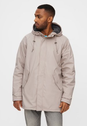 Mand iført en lysebeige vinterjakke med hætte og knaplukning på lommerne, kigger mod venstre foran en ensfarvet baggrund.