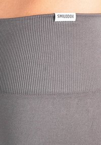 Grijze sportleggings met een geribbelde tailleband. Het materiaal lijkt rekbaar en glad, met een klein wit label bovenaan met de tekst "SMILODOX."