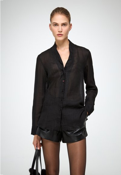Camicia nera trasparente a maniche lunghe con collarino, bottoni frontali e una sottile texture, abbinata a pantaloni corti in ecopelle nera.