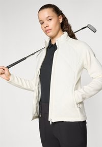 Leichte weiße Golfjacke mit geripptem Muster, Reißverschluss und Seitentaschen, getragen über einem schwarzen Poloshirt, kombiniert mit dunklen Hosen.