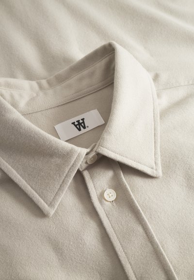 Chemise beige clair à boutons avec une finition texturée, dotée d'un col pointu, de boutons blancs et d'une étiquette de marque sur le col.