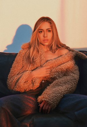 Ung kvinde med langt blondt hår iført en fluffy brun jakke og blå jeans, siddende på en mørk sofa med varmt lys.