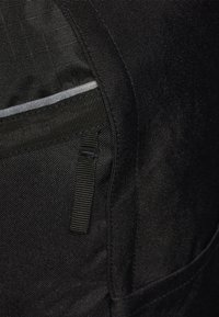 Svart tygväska med texturerad yta, som har en grå accentremsa och justerbar webbingrem för stängning. Inga synliga mönster eller logotyper.