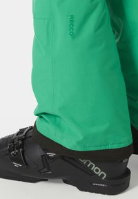 Jasně zelené lyžařské kalhoty s vyztuženými švy, logem Recco a černými akcenty. Obsahuje detailní záběr černé lyžařské boty s reflexními detaily.