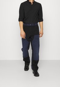 Camicia nera con bottoni abbinata a pantaloni in denim blu e scuro, caratterizzati da texture contrastanti e tasche angolate. Scarpe da ginnastica nere completano l'outfit.