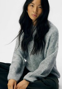 Grauer Wollpullover mit lockerer Passform, überschnittenen Schultern und gerippten Bündchen, kombiniert mit dunklen, fließenden Hosen. Schlichte Textur, minimalistisches Design.