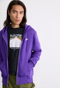 Corta-vento roxo com fecho éclair, apresentando um logótipo branco da Nike no peito, usado sobre uma t-shirt gráfica preta com um design de paisagem. Tecido texturizado com bolsos.