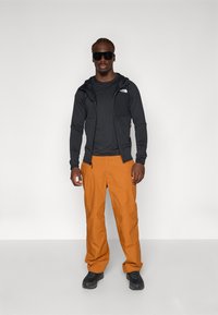 Haut zippé noir en tissu texturé, superposé à une chemise noire. Pantalons orange amples. Baskets noires et lunettes de soleil.