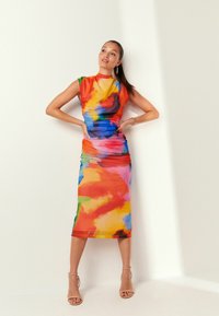 Eine Frau trägt ein ärmelloses, hochgeschnittenes, figurbetontes Midikleid mit einem mehrfarbigen abstrakten Muster und steht mit auf die Hüften gestützten Händen vor einer weißen Wand.
