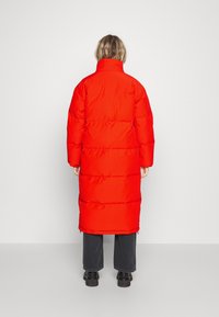 Rode pufferjas met een hoog collabel, voorzien van gequiltte secties en een lang ontwerp, gecombineerd met donkere denim broek en zwarte laarzen.