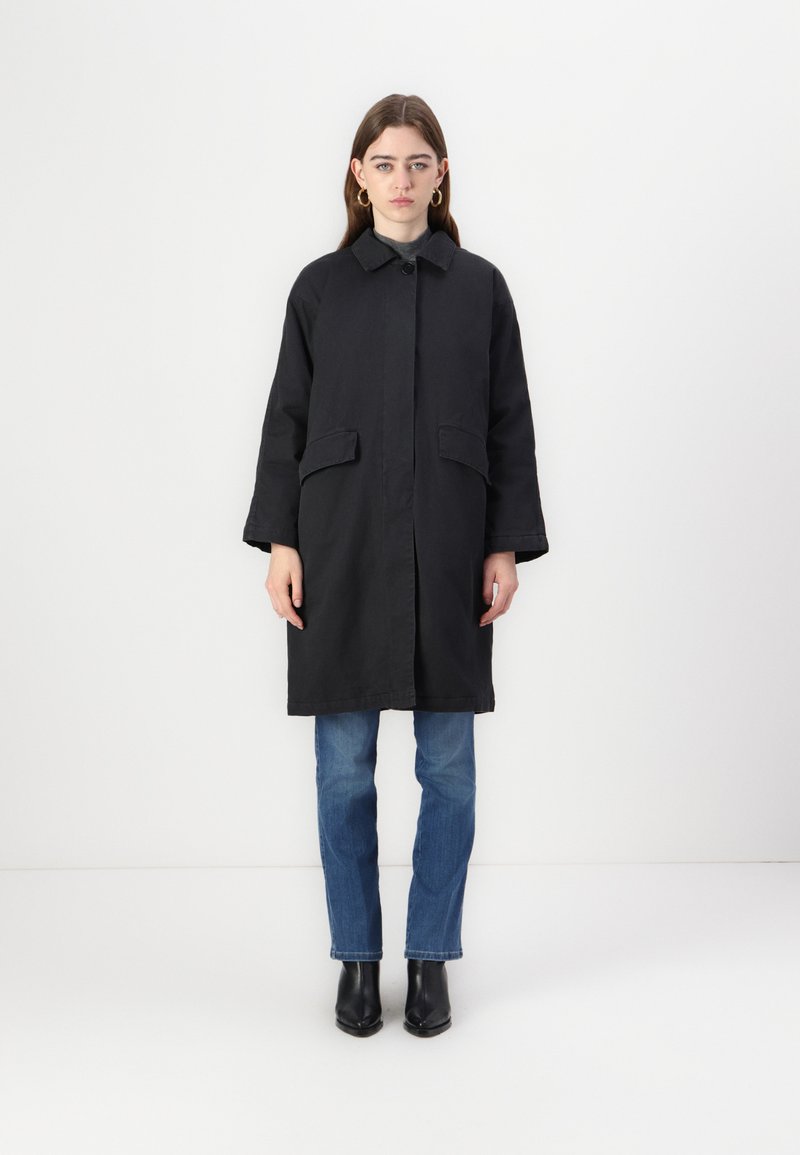 Black outerwear coat with a collared neckline, side pockets, and long sleeves. Worn over blue jeans and black ankle boots. Minimalist design.