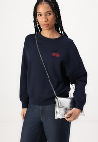 Námornícky modrý sweatshirt s červenou výšivkou "UŽÍVAJ SI DNES", skombinovaný s tmavými džínami s vysokým pásom a striebornou metalickou kabelkou.