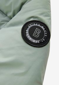 Svetlozelená prešívaná bunda s okrúhlou čiernou nášivkou s bielym textom a logom "B", vyrobená z hladkého, štruktúrovaného materiálu.
