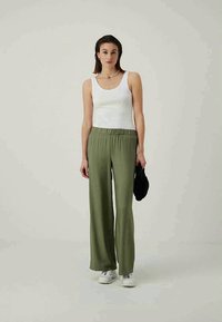 White tank top with wide green pants, featuring an elastic waistband. Model wears white sneakers and holds a black accessory. Plain background.