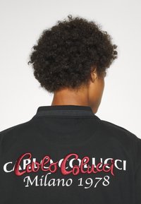 T-shirt polo noir avec texte brodé rouge et blanc au dos : "Carlos Colucci Milano 1978." Col détaillé avec motif géométrique.