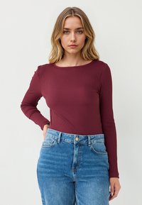 Burgunderfarbener gerippter Langarm-Top kombiniert mit hochgeschnittenen hellblauen Jeans. Das Top hat einen runden Ausschnitt, eine enganliegende Silhouette und eine weiche Textur.