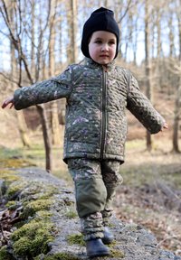 Tikitud rohelise jakiga, millel on lillemustrid, sobivad püksid ja mustad saapad. Laps kannab musta mütsi ja kõnnib samblal kaetud kivil metsas.