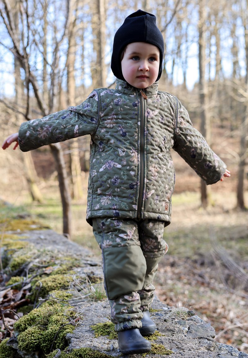 Tikitud rohelise jakiga, millel on lillemustrid, sobivad püksid ja mustad saapad. Laps kannab musta mütsi ja kõnnib samblal kaetud kivil metsas.