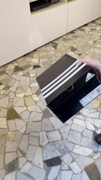 Boîte à chaussures noire avec une poignée sur le dessus, ornée de trois bandes blanches sur le couvercle et d'un logo adidas, posée sur un sol carrelé à motif.