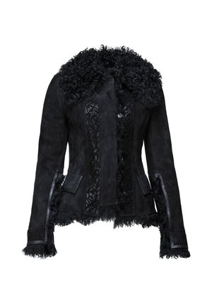 Femme en veste noire à col en fourrure, pantalon noir court et brillant, talons aiguilles noirs, tenant un petit sac noir avec chaîne dorée.
