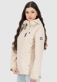 Giacca impermeabile beige con cappuccio, dotata di zip frontale, dettagli neri e due tasche laterali. Tessuto morbido e vestibilità rilassata.