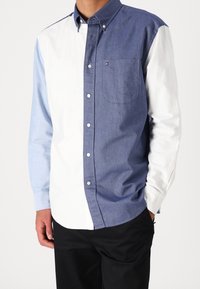 Camicia con bottoni caratterizzata da un design a blocchi di colore: pannelli in bianco solido, azzurro chiaro e denim scuro. Include una tasca sul petto e un colletto classico.