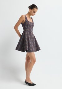 Femme avec les cheveux en chignon portant une robe sans manches à motif avec une jupe évasée et des chaussures à talons bas noires, se tenant devant un fond blanc uni.