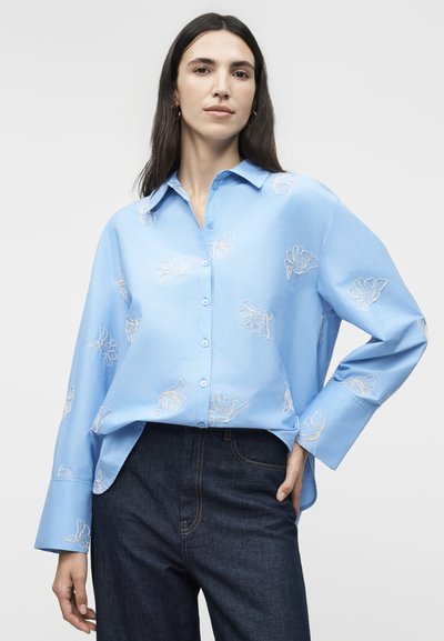 Γυναίκα που φοράει ανοιχτό μπλε πουκάμισο με λευκή κεντητή λουλουδάτη διακόσμηση και σκούρα μπλε τζιν, ποζάρει με το ένα χέρι στην τσέπη.