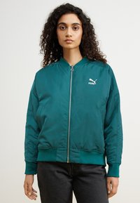 Tyrkis bomberjakke med lynlås foran, ribbede manchetter og kant, samt et lille hvidt Puma-logo på brystet. Glat, shiny stoftekstur.