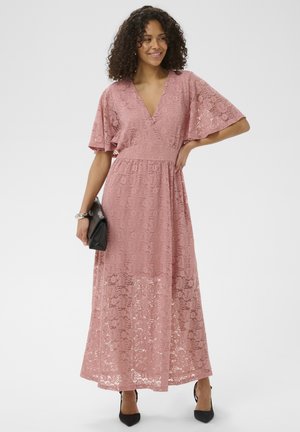 Donna con un abito lungo in pizzo rosa polvere con maniche svolazzanti, che tiene una pochette nera e indossa tacchi neri, sorridente e in posa.