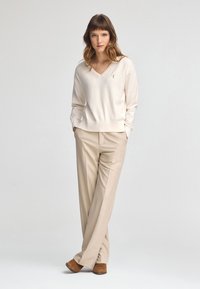 Maglione crema a V con orlo a coste, abbinato a pantaloni beige chiaro. Il modello è in piedi con le mani in tasca, indossando scarpe color nocciola.