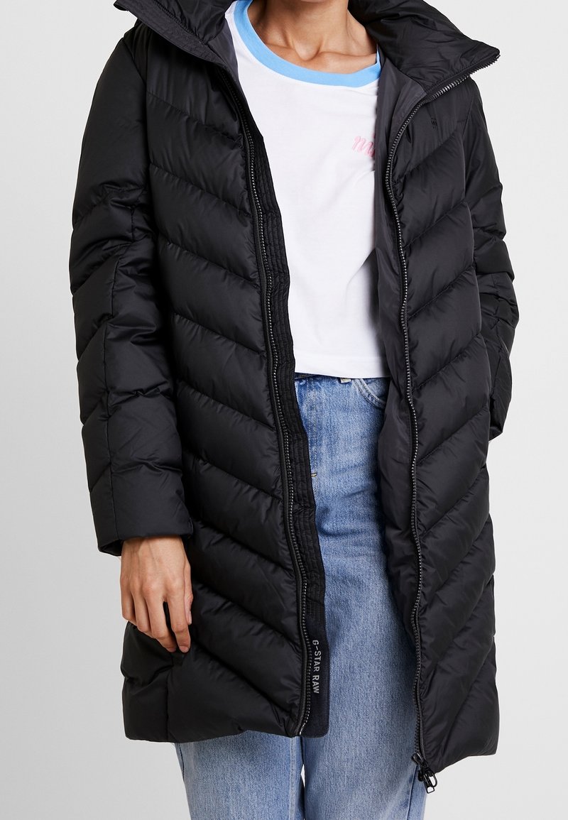 Zwarte pufferjas met capuchon, voorzien van horizontale doorgestikte panelen, ritssluiting en een lange pasvorm. Draagt over een wit T-shirt en een spijkerbroek.