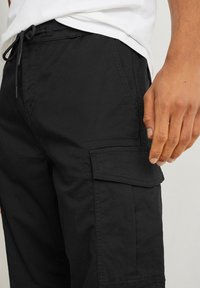 Gros plan sur une personne portant un pantalon cargo noir avec un cordon de serrage à l'avant et une poche à rabat sur la cuisse, la main reposant à côté de la poche.