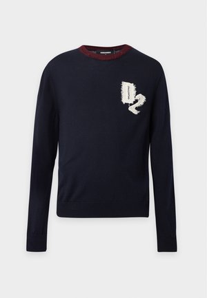 Jeune homme portant un pull noir avec un patch "D2", un jean skinny gris clair déchiré et des baskets noires à rayures blanches, debout sur un fond blanc.