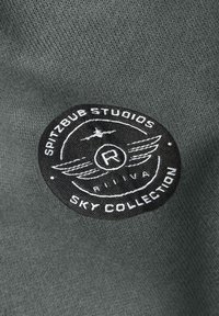 Zwarte, ronde stoffen patch met witte tekst "Spitzbub Studios Sky Collection" en een gevleugeld embleem op donkergrijze gebreide stof.