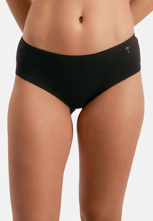 Naisen vyötärö ja yläreidet, joilla on yksiväriset mustat hipsterialushousut, joissa on pieni kultainen tunnus vasemmalla puolella.