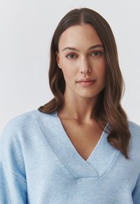 Lyseblå V-hals sweater lavet af blødt stof, der har en afslappet pasform og ribbede detaljer ved halsudskæringen. Den strukturerede overflade er synlig.