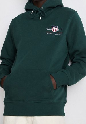 Person trägt dunkelgrünen GANT-Hoodie mit Vordertasche und Logo-Emblem auf der Brust, Hände teilweise in der Tasche, weiße Hose sichtbar.