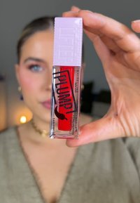 Produit pour les lèvres dans un contenant transparent avec une teinte rouge, doté d'un capuchon violet texturé. Le texte sur les étiquettes inclut "PLUMP" et "MAYBELLINE."
