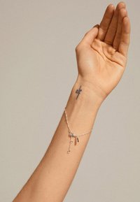 Sidabrinė grandinėlė su mažos mėlynos brangakmenio ir raktų formos akcentais, dėvima ant rankos su matoma odos tekstūra ir tatuiruote.