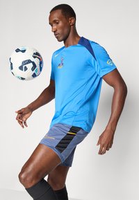 Zila sporta krekls ar logo, īsi pelēki šorti ar melnu svītru un dzeltenu akcentu, melnas zeķes, un futbola bumba gaisā.