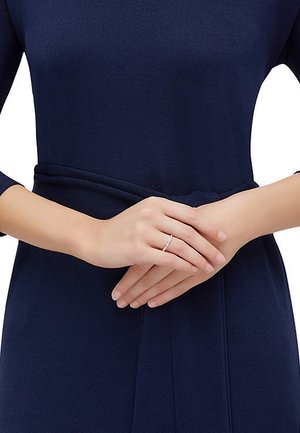 Silberring, der an den Fingern getragen wird, schlankes Design mit einer glatten Oberfläche, über einem dunklen marineblauen Stoffkleidungsstück, das die Hände betont.