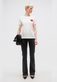 Γυναίκα με κοντά ξανθά μαλλιά, φορώντας λευκό T-shirt με κόκκινη καρδιά, μαύρο τζιν παντελόνι, μαύρα παπούτσια, κρατώντας μαύρο μπουφάν.