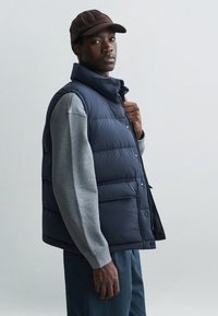 Chaleco acolchado azul marino con cuello alto, cierre de botones y bolsillos laterales, combinado con una camiseta de manga larga gris. Una gorra oscura completa el look.
