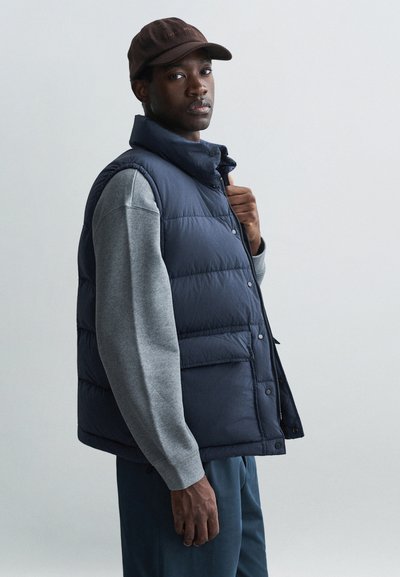 Marineblauwe puffervest met een staande kraag, knoopsluiting en zijzakken, gecombineerd met een grijs shirt met lange mouwen. Een donkere pet maakt de look compleet.